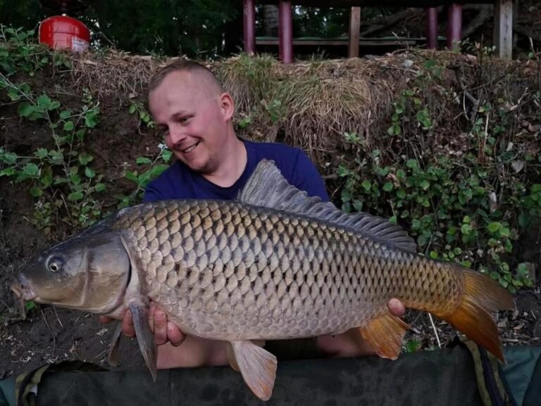 Michal kľačí na brehu s kaprom uloveným na boilies Anaconda AI Carp Shellfish & Plum 24 mm